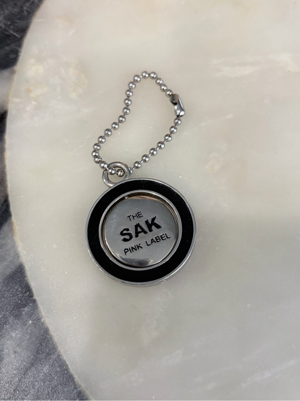 The Sak Silver and Black Round bag Sharm/ Logo Keychain